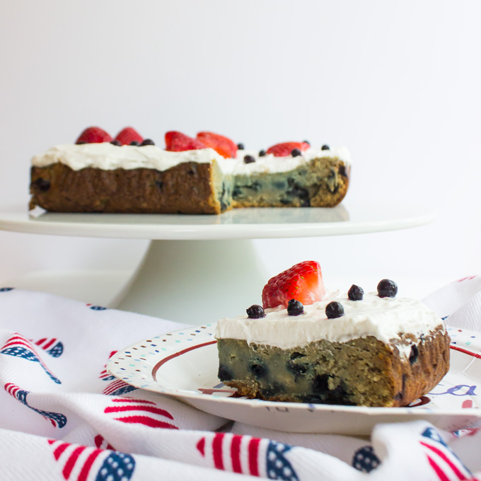 Side view of a slice of Blueberry Snack Cake with strawberry and blueberry toppings placed in a USA flag themed paperplate over a USA themed mantel with a sliced blueberry snack cake placed in a cake stand round plate in the background