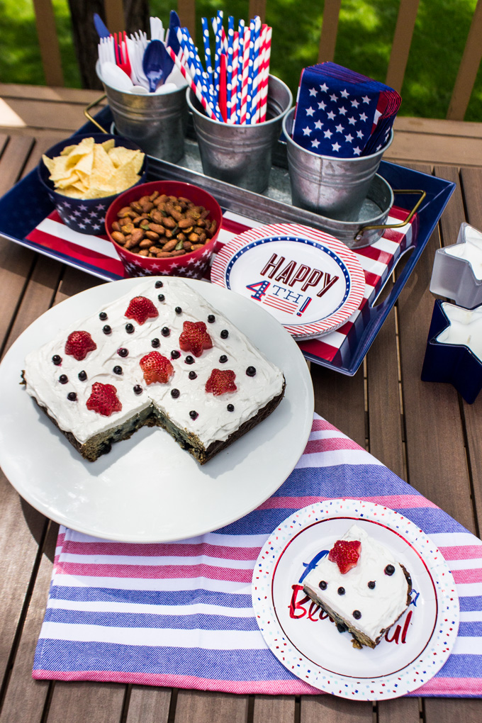 4th of July themed portrait of a sliced blueberry snack cake with strawberry and blueberry toppings in a white round plate alongside a slice of the cake in a paperplate and a tray of 4th of July paperplate, bowl of almond and potato chips, and a tray of 3 aluminum ice bucket filled with USA flag themed napkins, straw, and plastic cutlery.