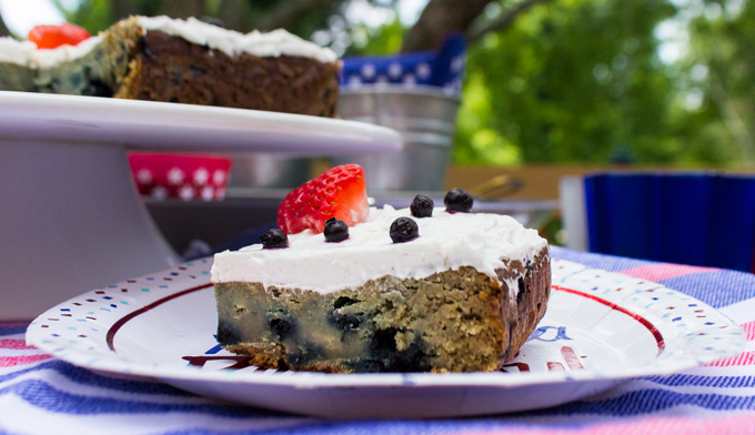 Side view of a slice of blueberry snack cake with strawberry and blueberry toppings placed in a 4th of July themed paperplate and over a USA flag themed mantle