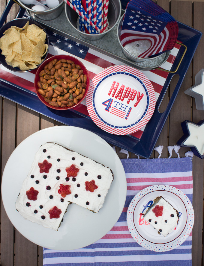 Overhead view of Blueberry Snack Cake with a bowl of almond, potato chips, Happy 4th of July plate, 3 tray of aluminum ice bucket with USA flag, USA flag themed straw, and a plastic spoon all in a blue tray with a slice of Blueberry Snack Cake in a dessert plate beside it
